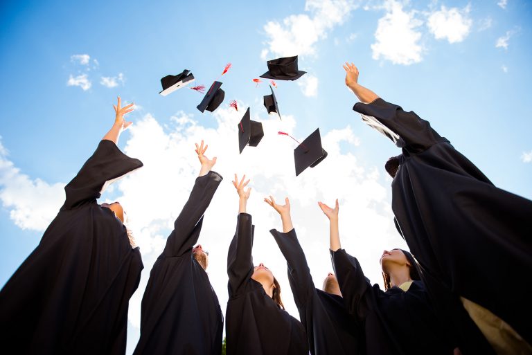 Happy group of six young graduates in black gowns, throwing up their head wear in the ai